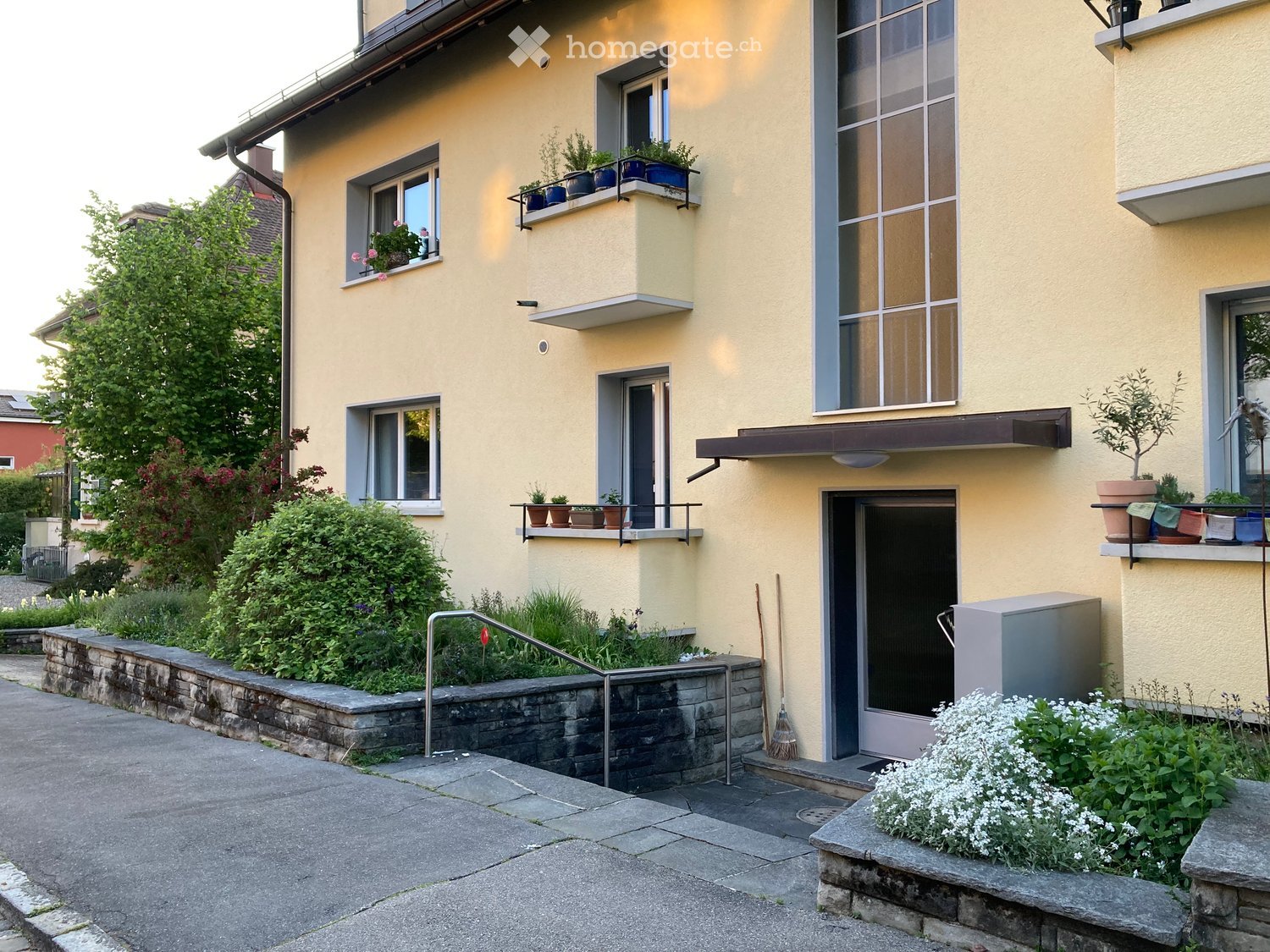 Two-story yellow building with multiple windows, balconies, and potted plants. Features a raised ground floor with a stone retaining wall and greenery.