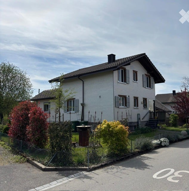 Two-story house with white exterior walls, multiple windows, and a dark roof. The house is surrounded by a small garden with various plants and trees.
