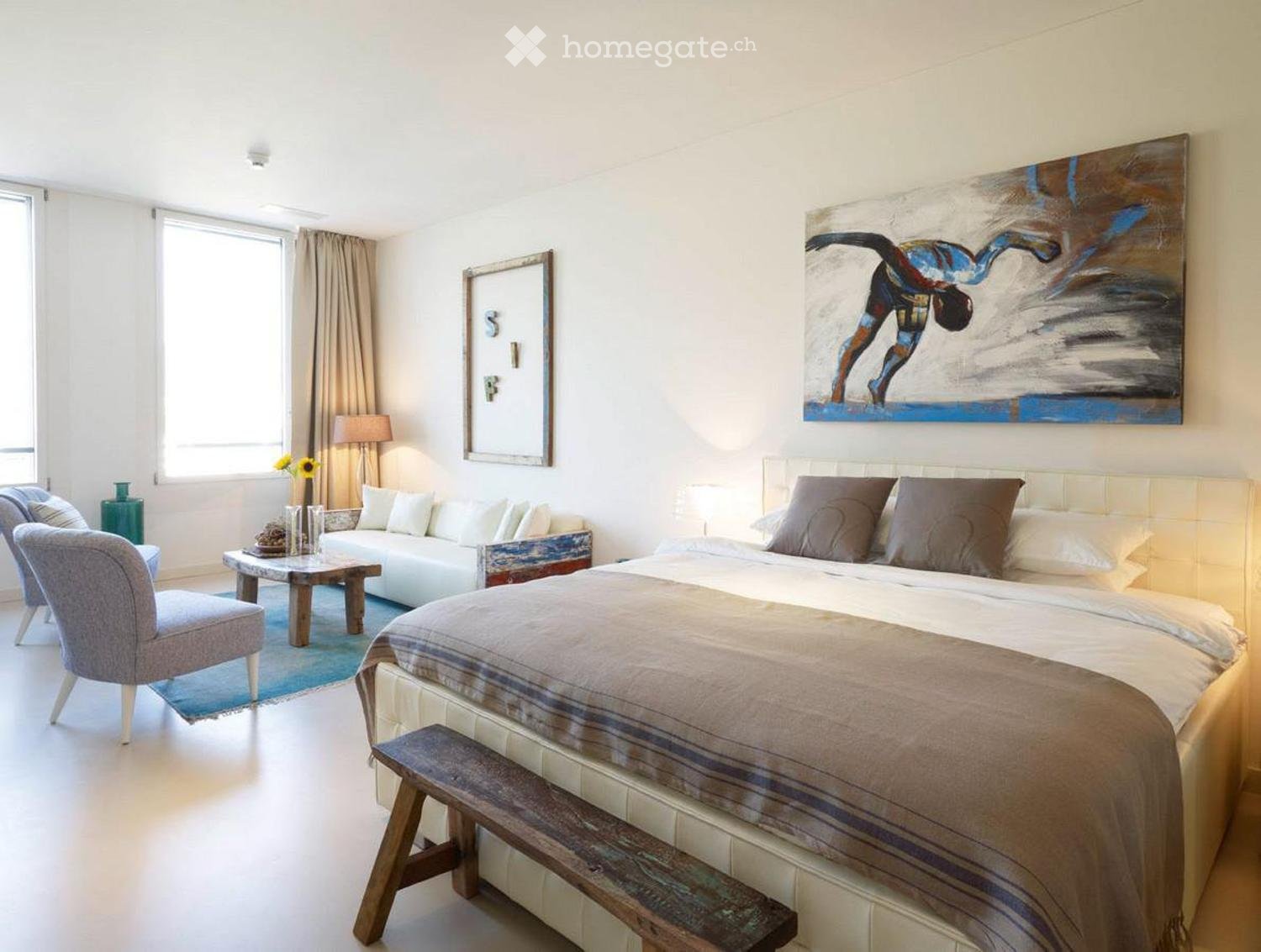 Bright bedroom with a large bed, sofa, wooden chair, small table, and artwork on the walls.