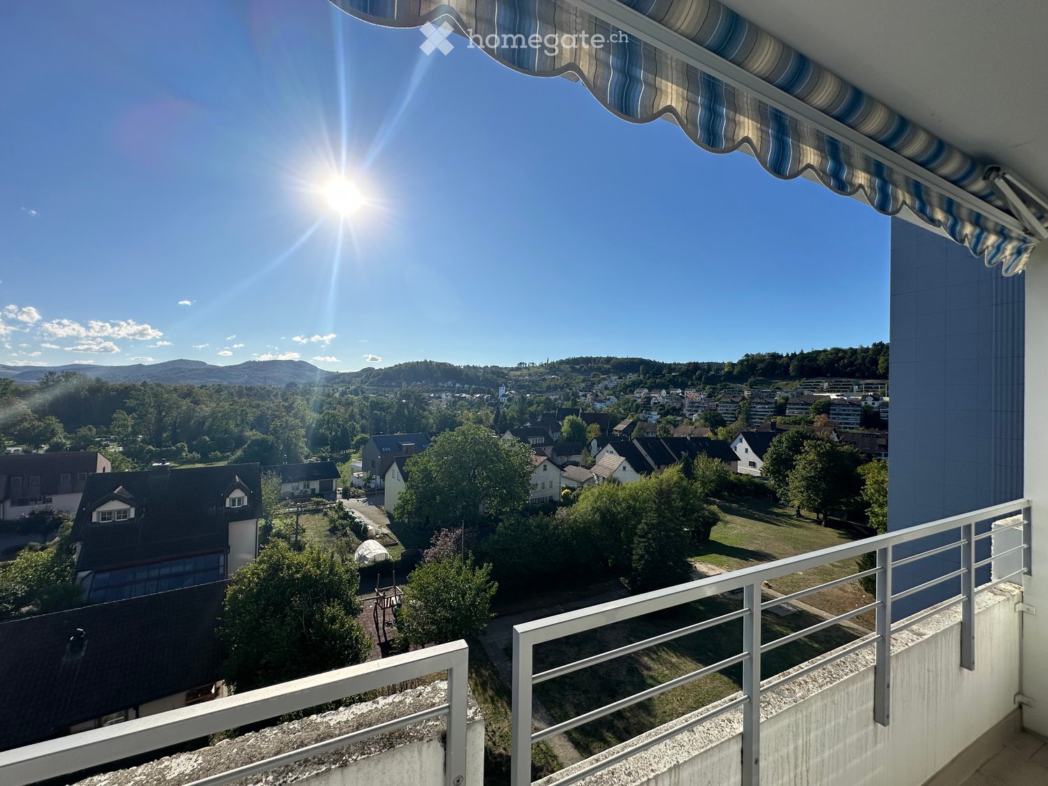 Balcony with a view of a town and surrounding landscape, featuring a striped awning and metal railing.