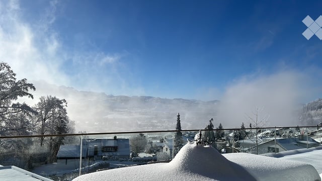 A snowy balcony or terrace overlooking a mountain landscape with a town below, clear blue sky, and snow-covered roofs.