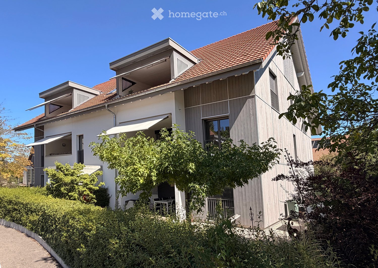 Modern two-story house with a red-tiled roof, white and grey exterior, multiple windows, and a balcony on the upper level. The house is surrounded by greenery and has a well-maintained garden.