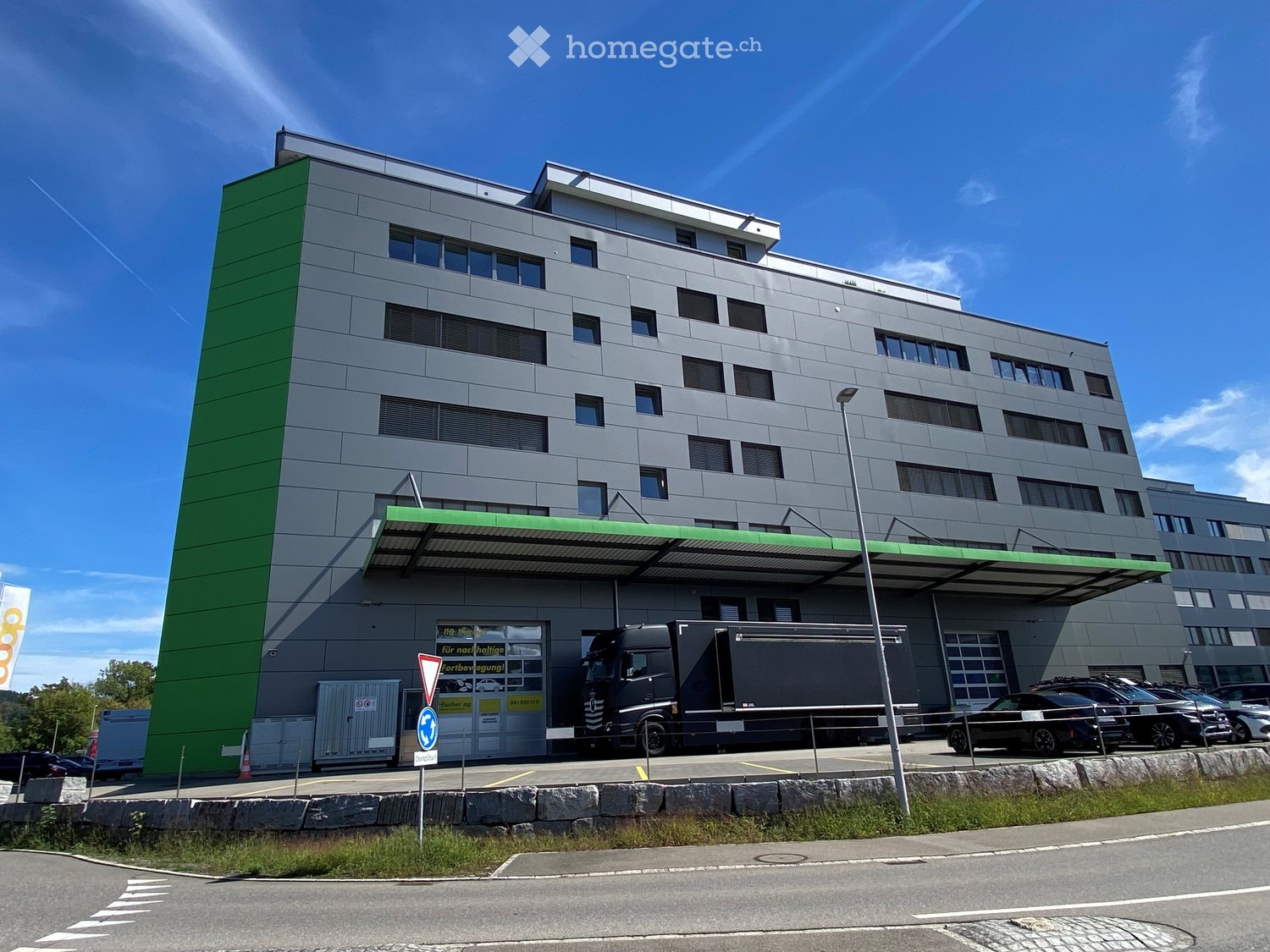 Modern multi-story building with grey facade and green accents, featuring a large truck parked in front and several cars in the parking area.