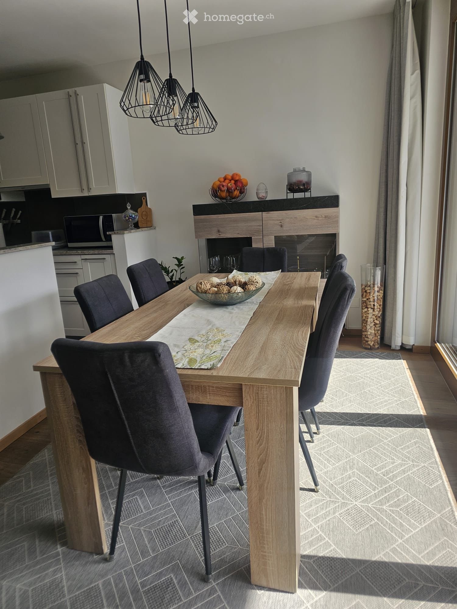 Modern dining area with a wooden dining table, six dark grey chairs, a decorative bowl with decorative items, a fireplace, and pendant lights. The area is connected to a kitchen with white cabinets and a microwave.