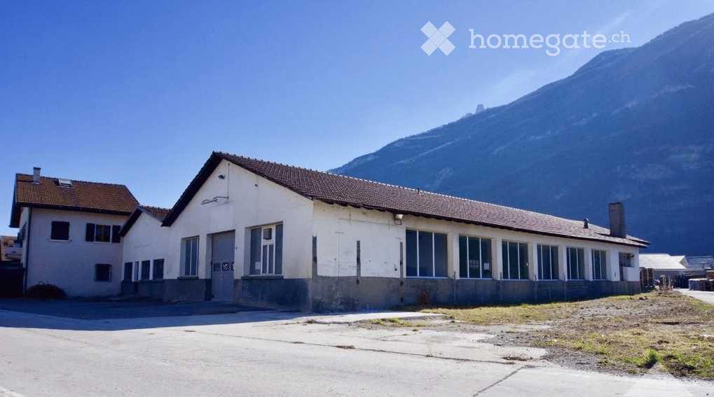 A two-story building with a flat roof and large windows, situated in a rural area with a mountain in the background.