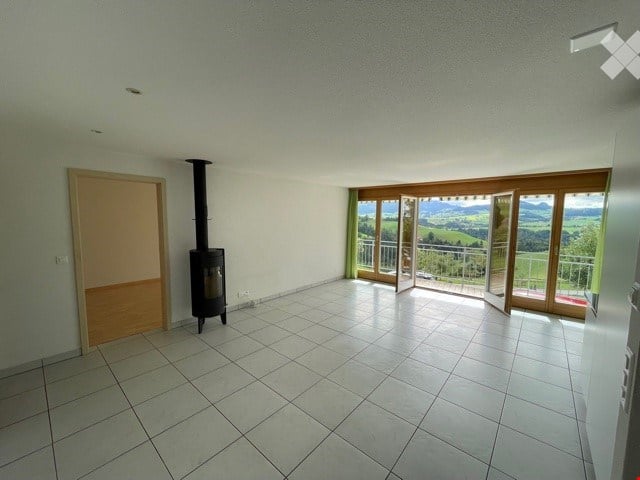 Empty room with tiled floor, white walls, a wood-burning stove, and large windows offering a scenic view of the landscape.