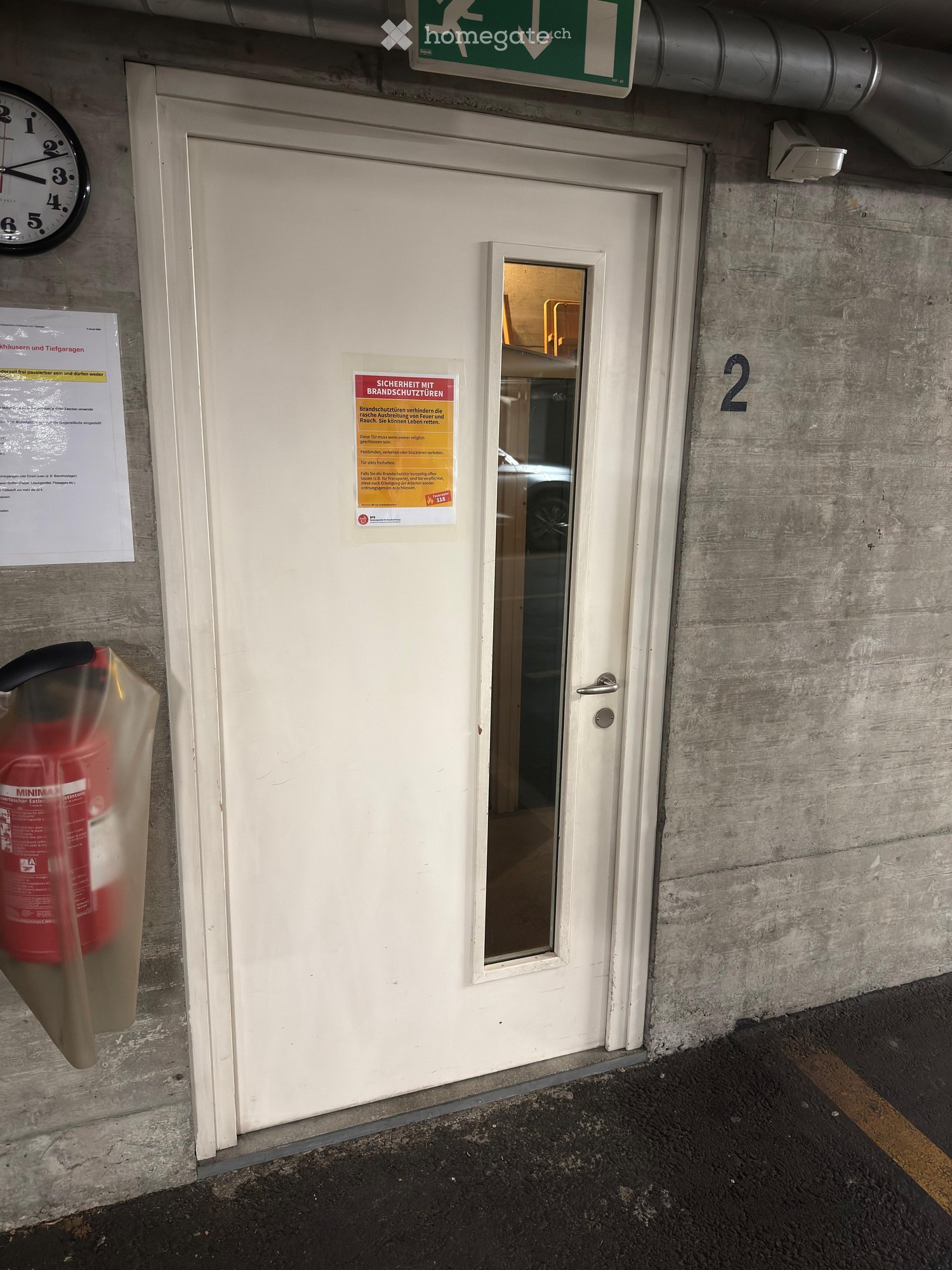 A white door with a small window, a fire extinguisher mounted on the wall, a clock, and a green emergency exit sign above the door.
