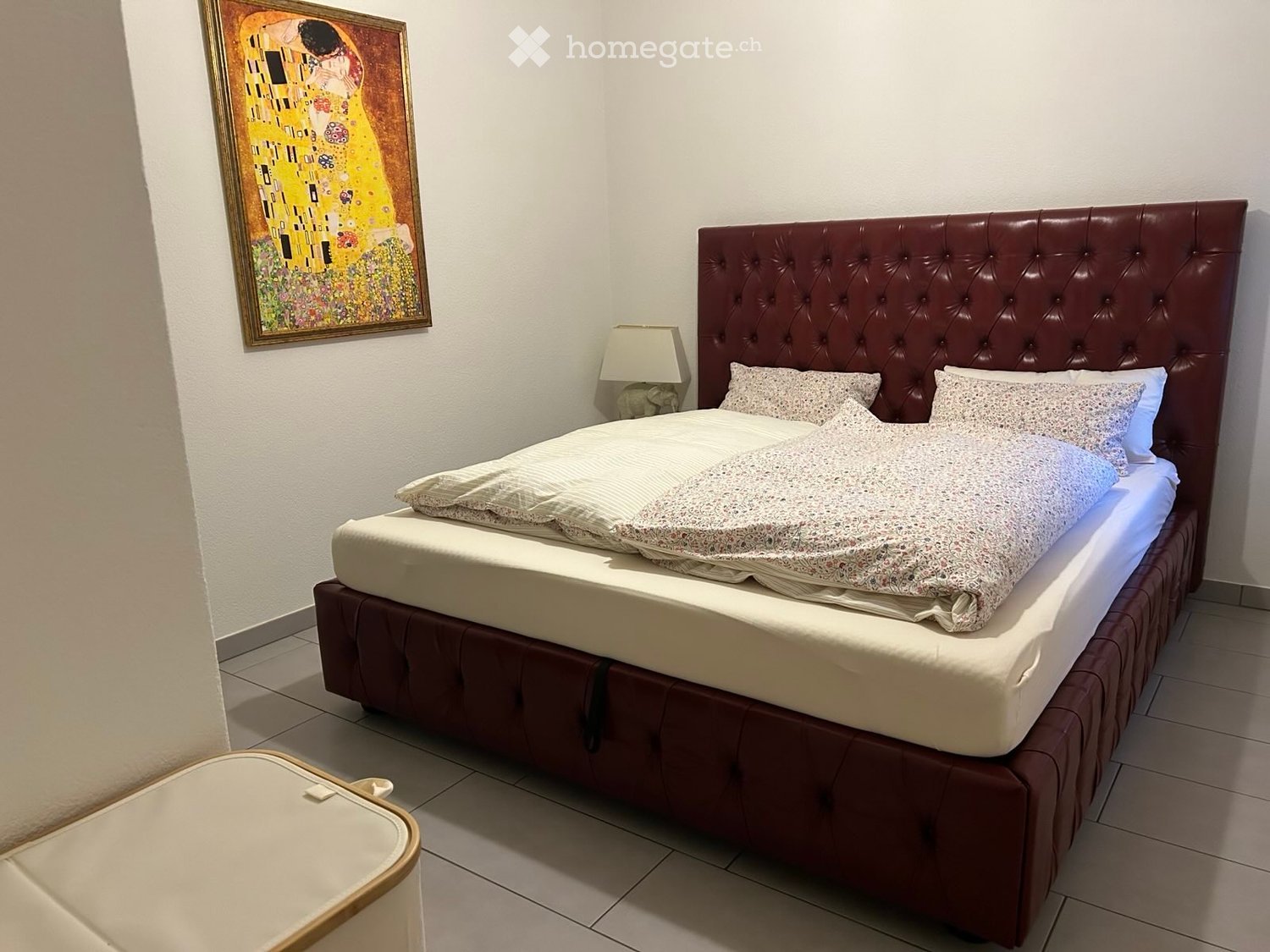Cozy bedroom with a large upholstered bed, patterned bedding, a nightstand with a lamp, and a framed painting on the wall.