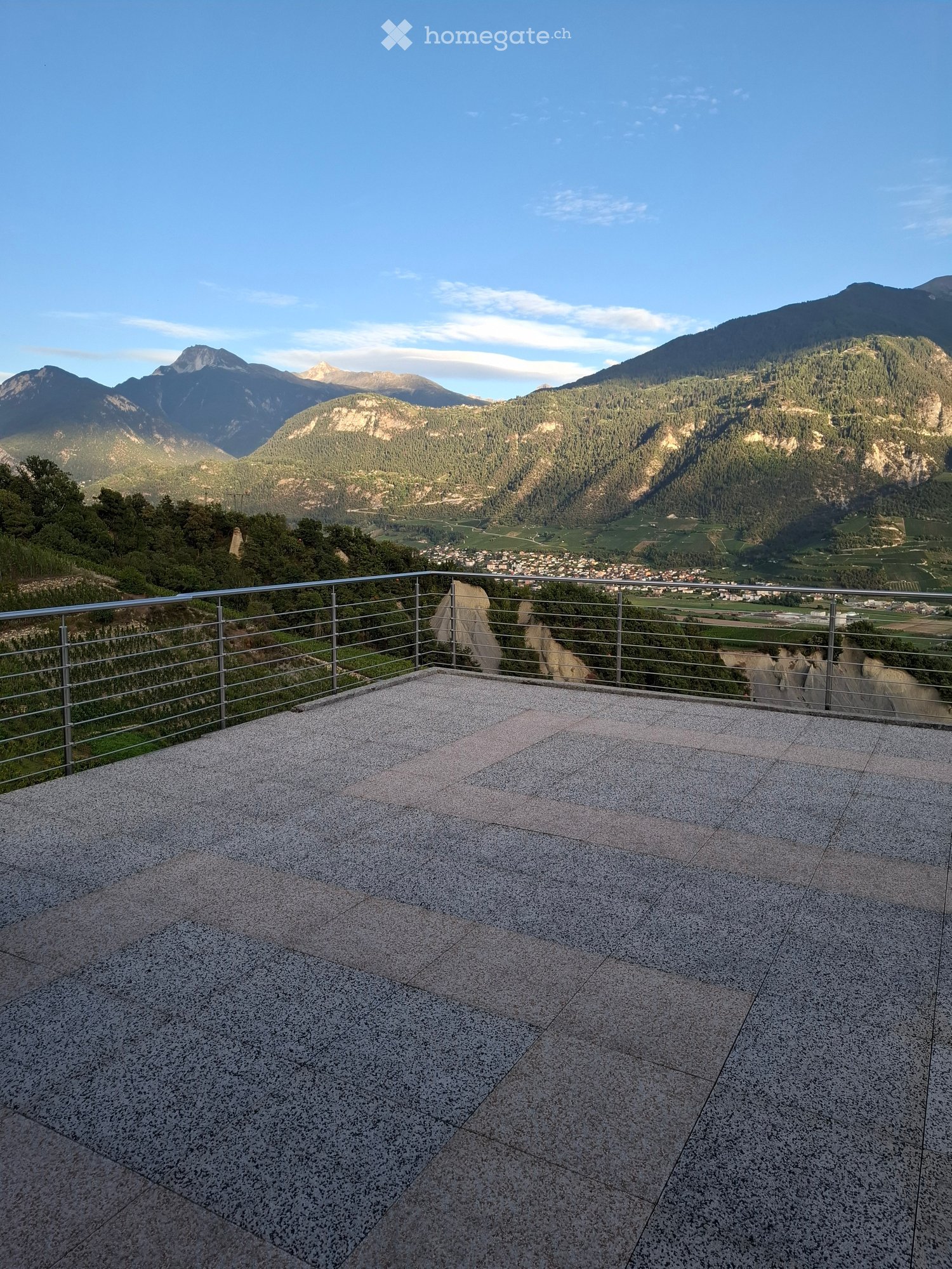 A spacious balcony with a modern metal railing, overlooking a scenic valley and mountains.