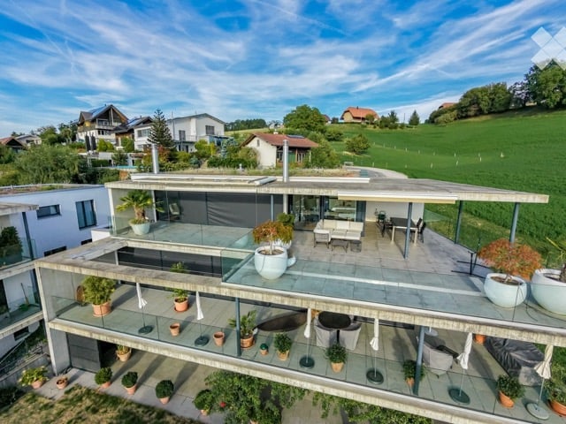 Modern two-story house with a large terrace and glass railings, surrounded by greenery and potted plants. The house features a flat roof and contemporary design.