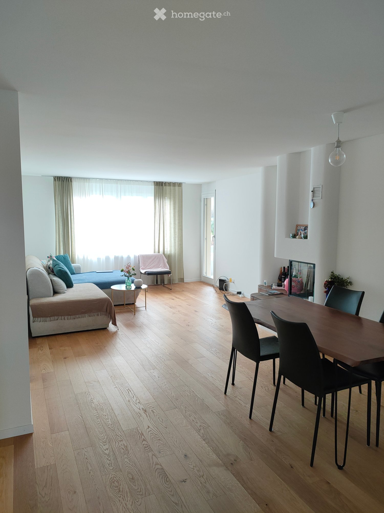 Open-plan living and dining area with light wooden flooring, a sofa, a small coffee table, a dining table with chairs, and a vase on a side table.