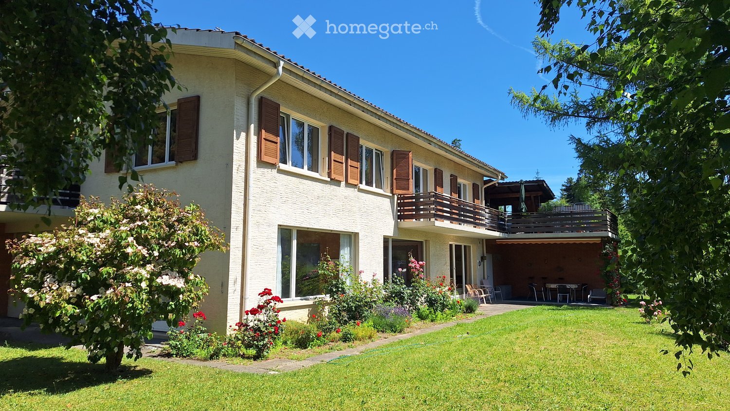 Two-story house with white exterior, wooden shutters, large windows, and a balcony. The house is surrounded by a well-maintained garden with green grass, flowering bushes, and trees.