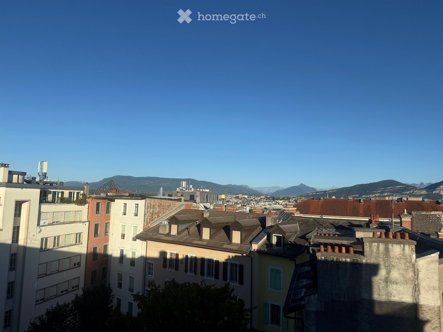 A cityscape view showing multiple buildings with varied roof styles and colors, set against a backdrop of distant mountains and a clear blue sky.