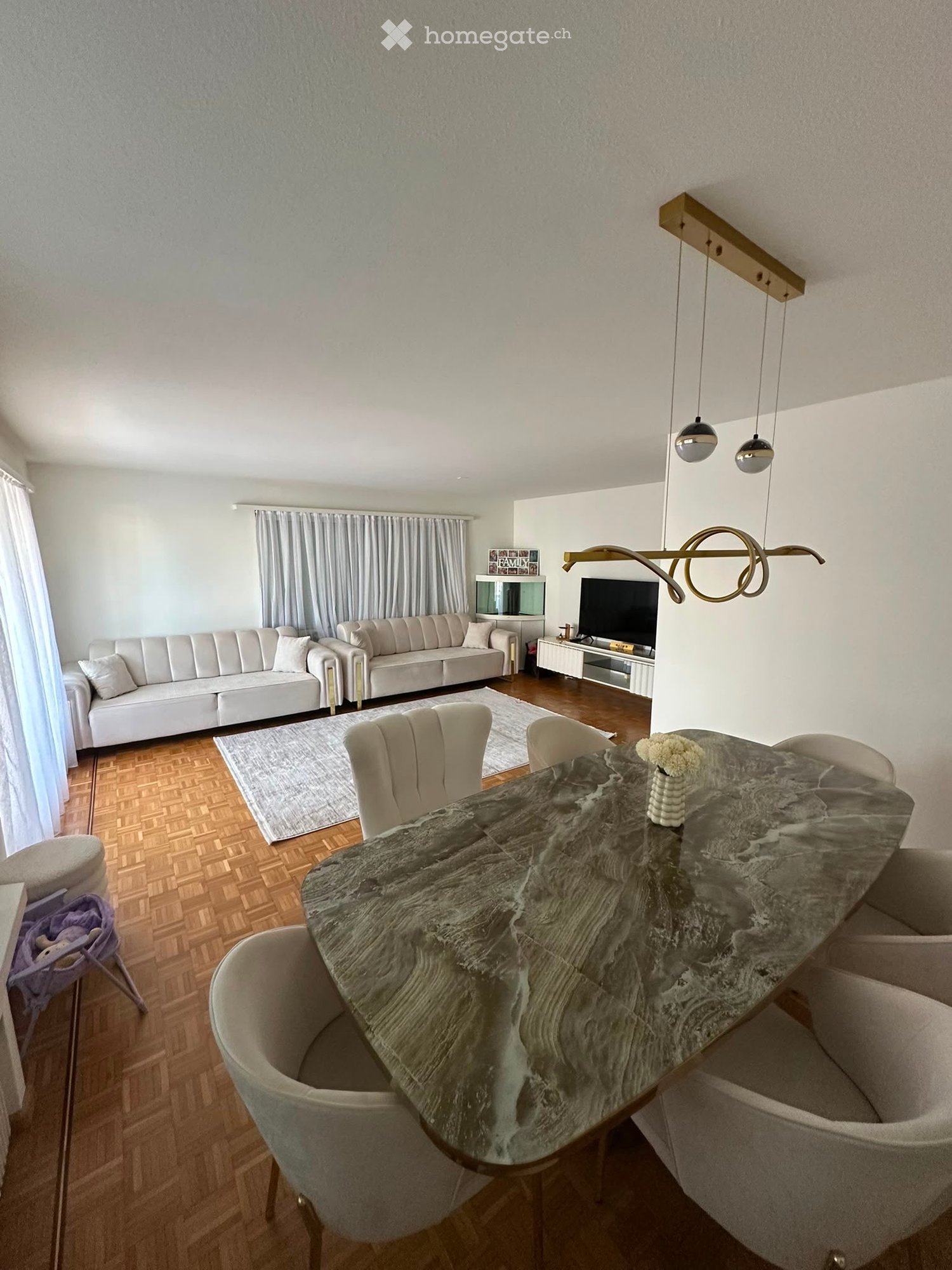 Modern living and dining area with a white sofa, grey dining table, chairs, and a television. The room features wooden flooring, a large window with curtains, and modern lighting fixtures.