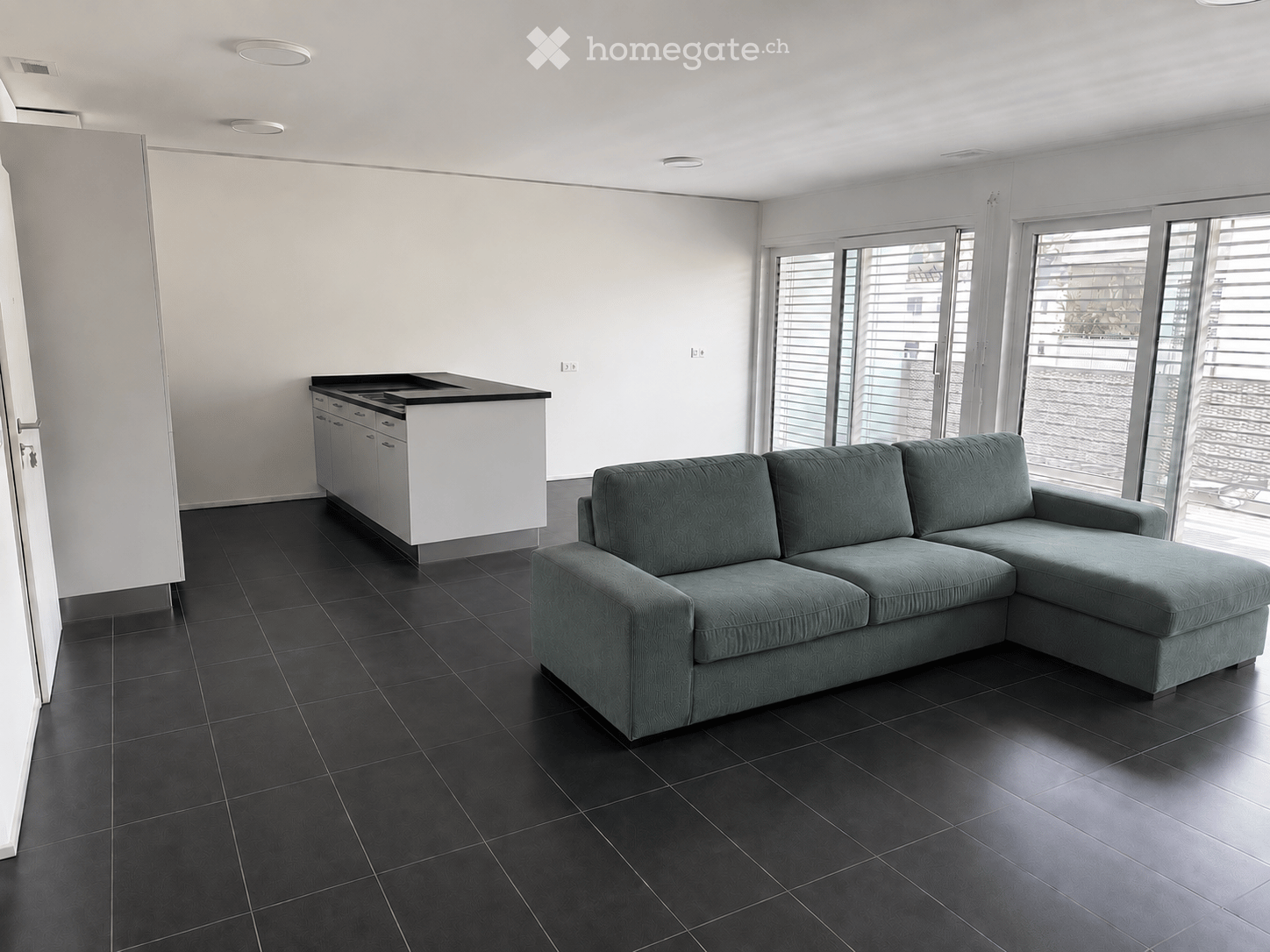 Modern living room with a grey sectional sofa, black tiled floor, white kitchen island with cabinets, and large windows with blinds.