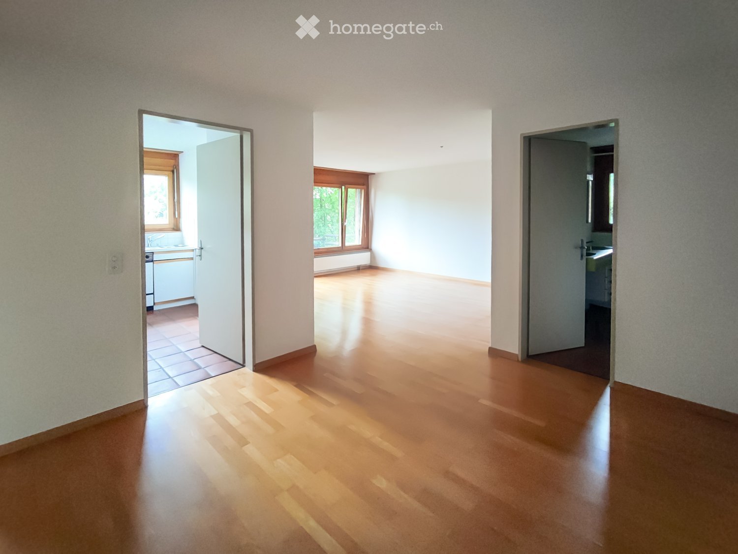 Empty room with wooden flooring and white walls, featuring open doors leading to a kitchen and bathroom.