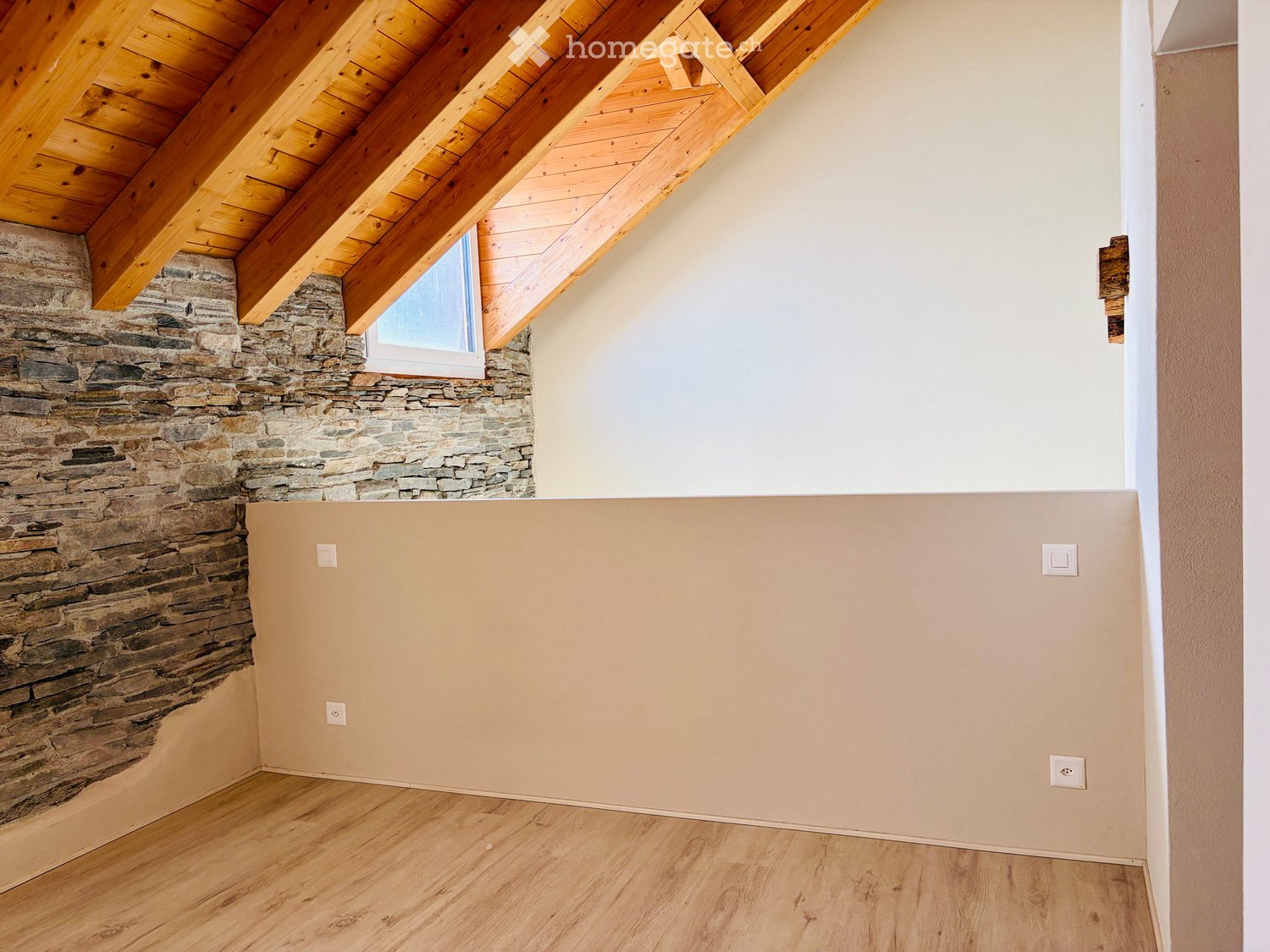 Empty room with wooden flooring, exposed wooden beams on the ceiling, and a stone accent wall. A small window is visible in the stone wall.