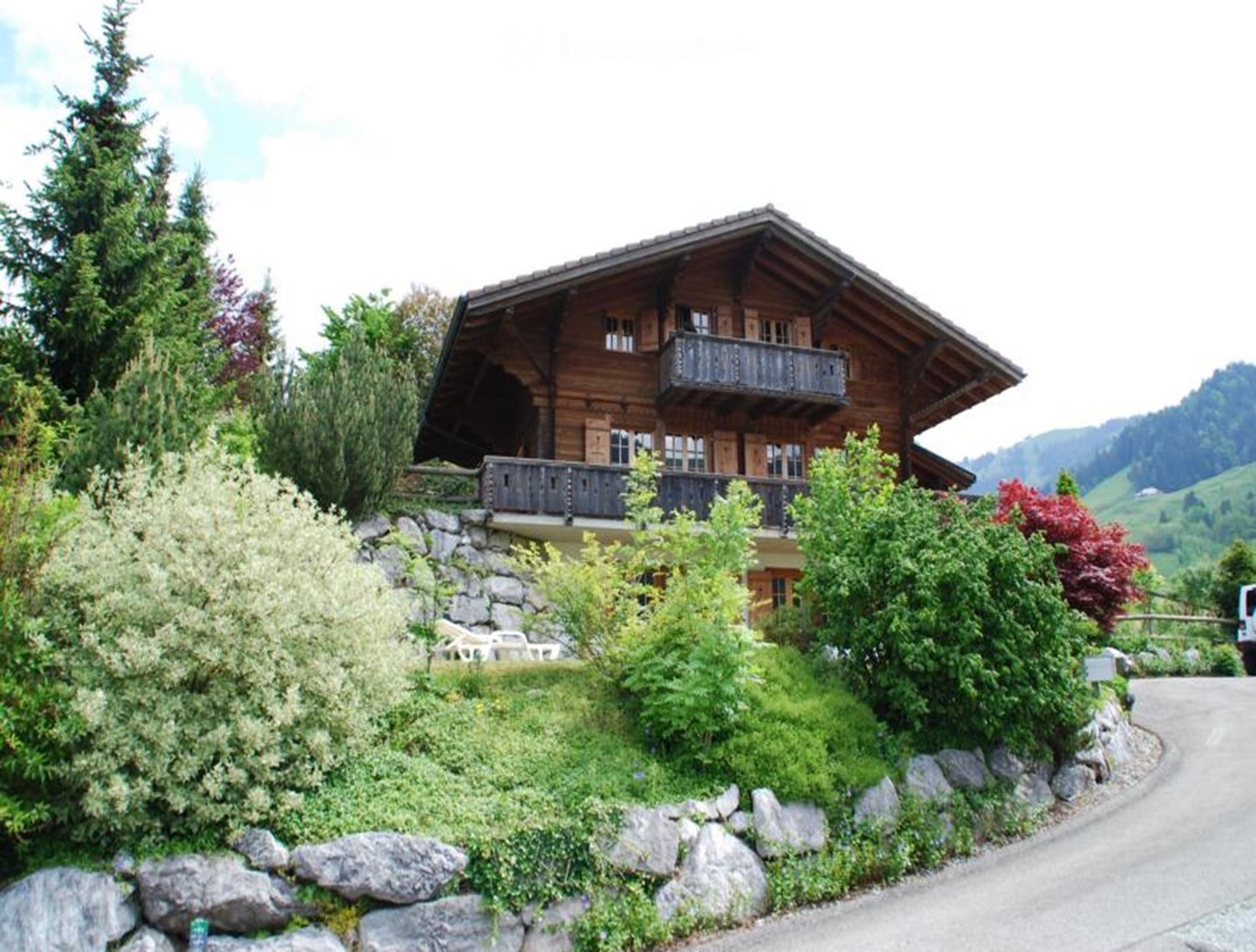 A two-story wooden house with balconies, surrounded by greenery and a stone retaining wall.