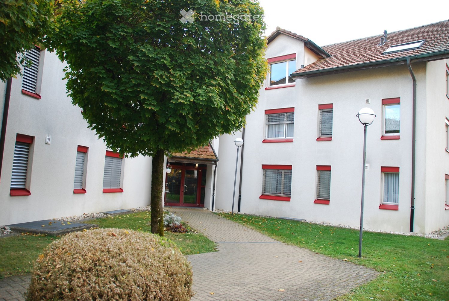 Two-story residential building with white walls, red window frames, and a tiled roof. The entrance features a red door and a small porch. The building is surrounded by a well-maintained lawn, a tree, and a paved pathway.