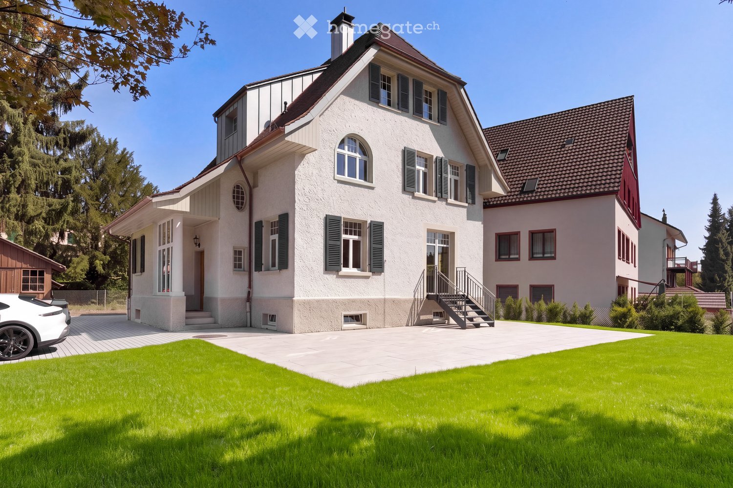 A two-story house with white walls, green shutters, and a well-maintained lawn in front. The house features a driveway, a staircase leading to the entrance, and a small wooden shed to the left.