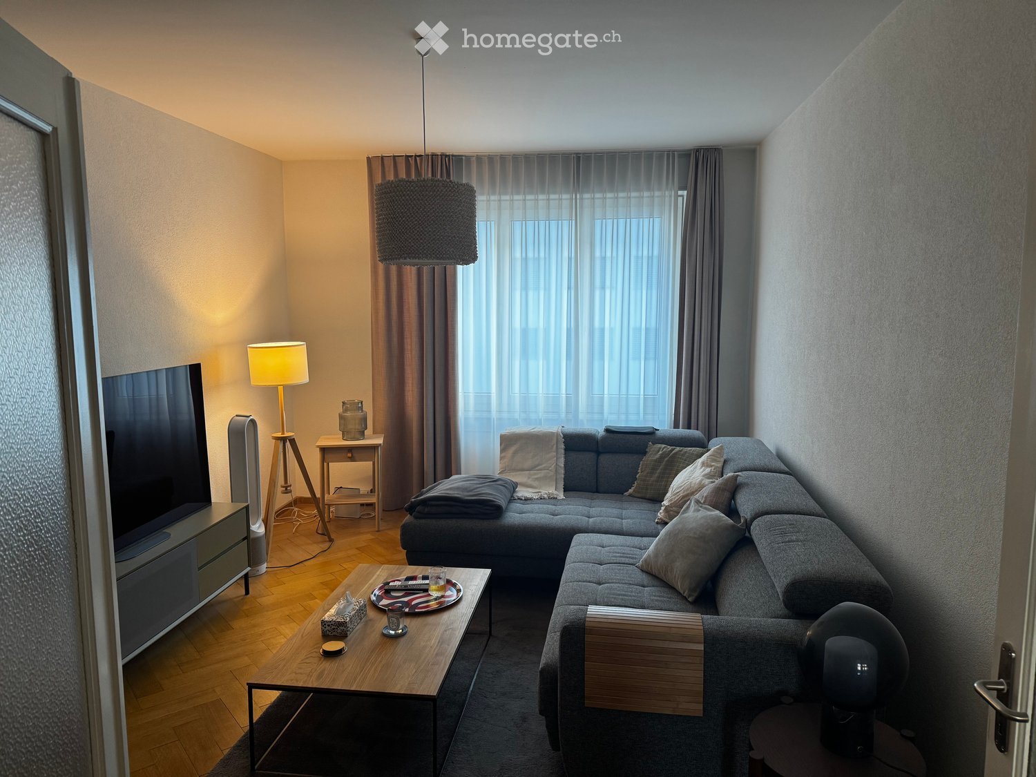 Modern living room with a grey sectional sofa, wooden coffee table, flat-screen TV, and warm lighting from table and floor lamps.