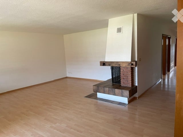 Empty room with wooden flooring, white walls, and a fireplace with a brick surround.