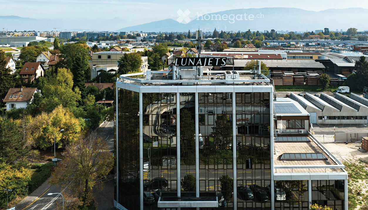 A modern, multi-story building with reflective glass windows and a rooftop sign reading "LUNAJETS." The structure is surrounded by trees, other buildings, and a parking area with several cars.