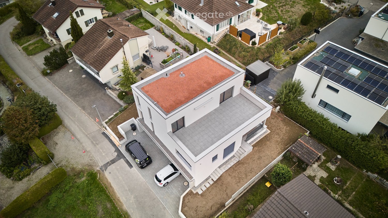 Aerial view of a modern white house with a flat roof, garage, and parking area. The house has large windows and a contemporary design. Solar panels are visible on neighboring roofs.