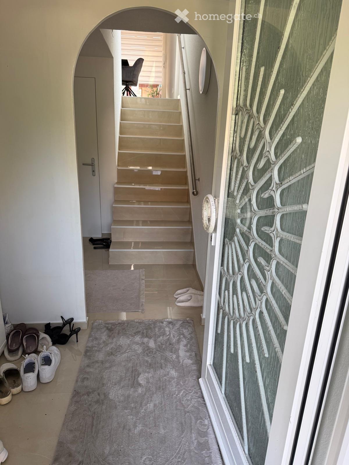 Hallway with a staircase leading upwards, a glass door with decorative pattern on the right, and a gray rug on the floor. Shoes and slippers are placed near the entrance.