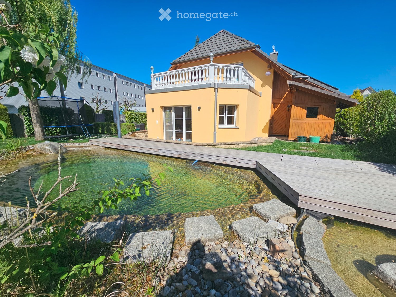 Garden with a small pond, wooden deck, stone steps, and a yellow house with a balcony.