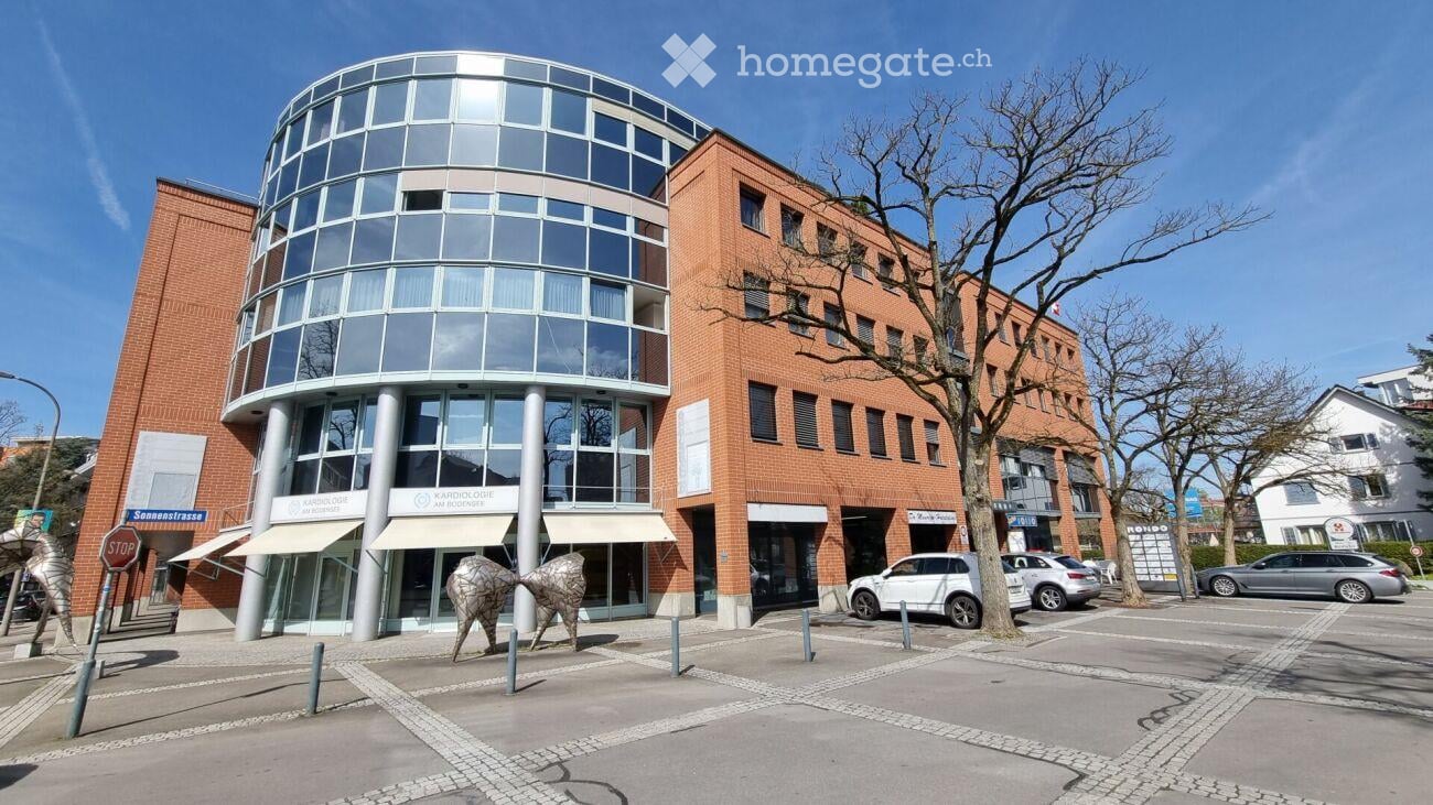 Modern multi-story building with a mix of red brick and glass facade, featuring a rounded glass tower section and several parked cars in front.