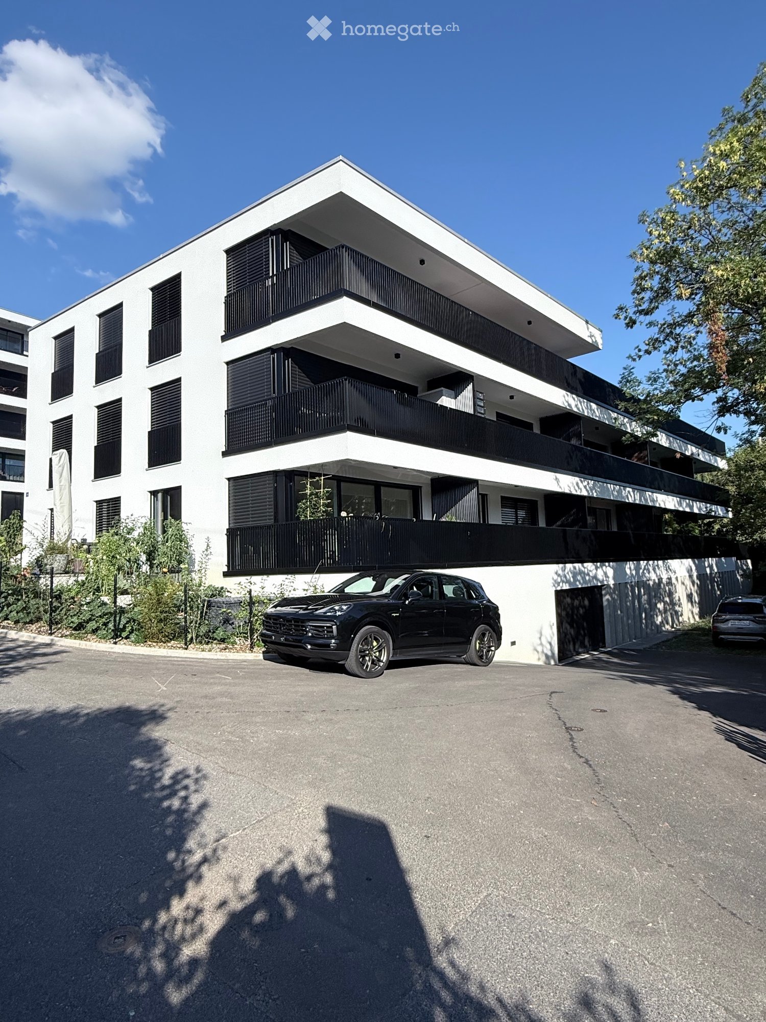 Modern three-story residential building with white facade, black balconies, and large windows. A black SUV is parked in front of the building.