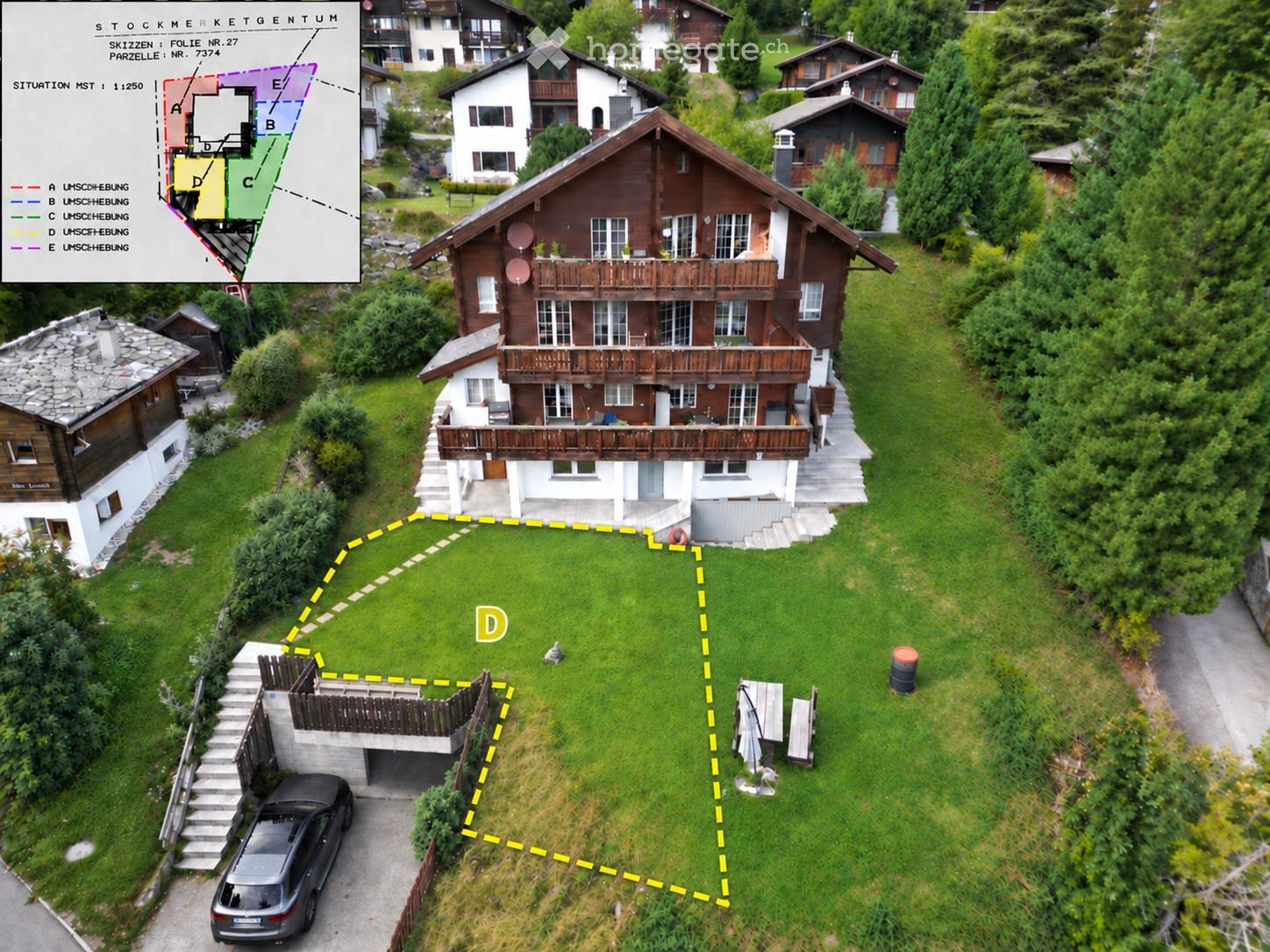 Aerial view of a two-story wooden house with a large balcony, surrounded by a well-maintained lawn and neighboring houses. A car is parked in the driveway, and a small garden area is visible.