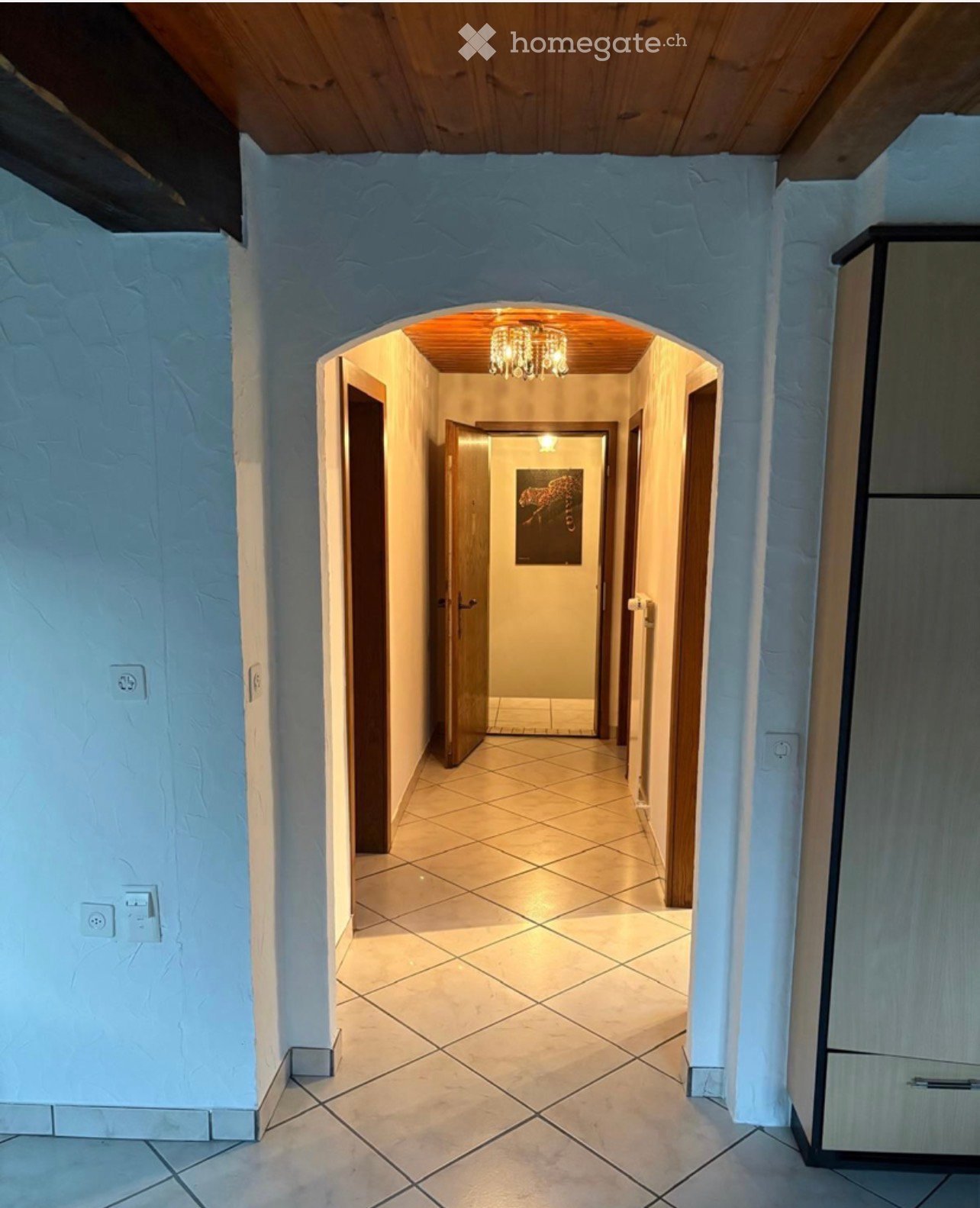 A well-lit hallway with tiled flooring, light blue textured walls, and wooden doors leading to other rooms. A chandelier hangs from the ceiling at the far end.