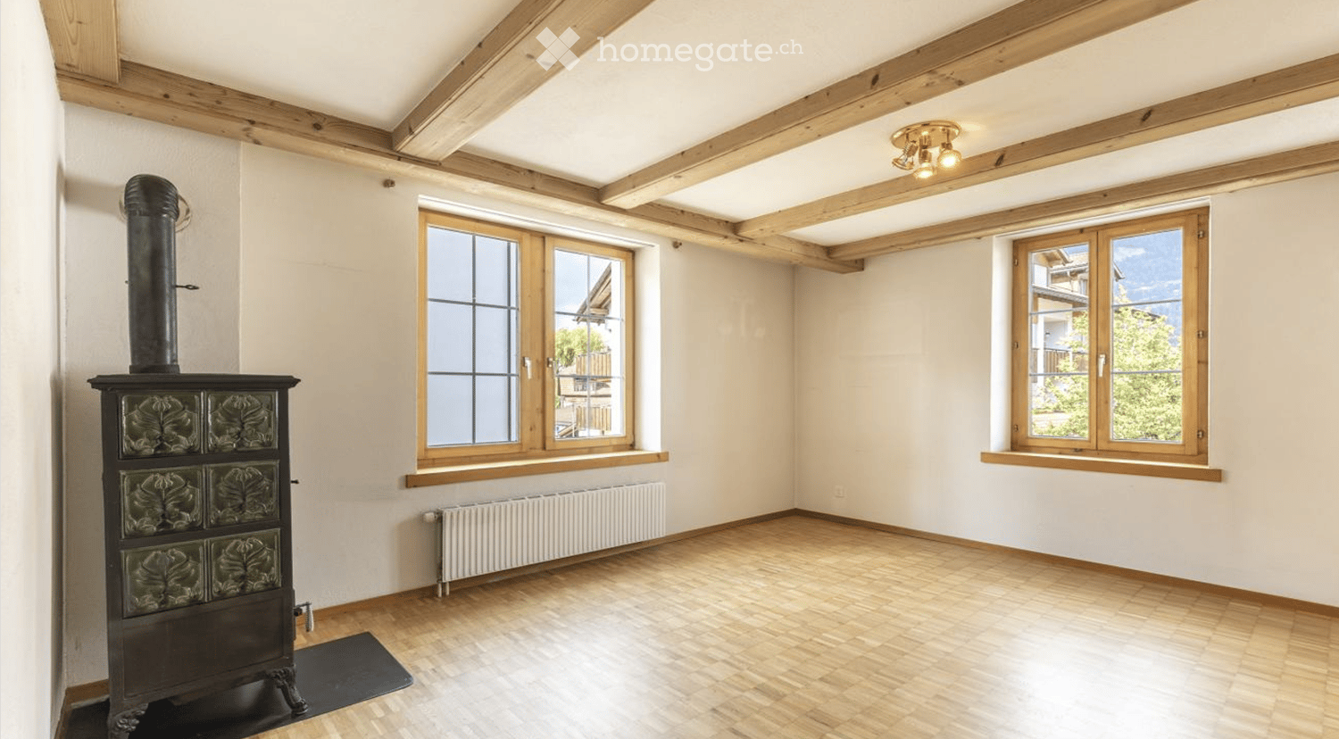 Empty room with wooden floor, exposed wooden beams on the ceiling, white walls, two large windows, radiator, and a decorative stove with a chimney pipe.