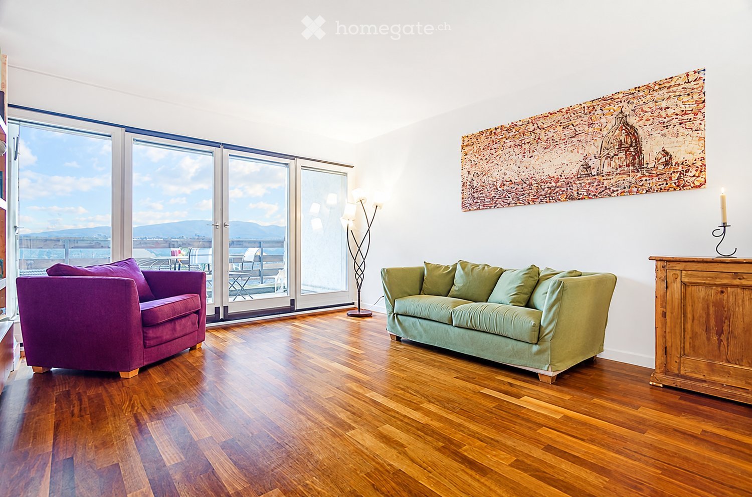 Modern living room with wooden flooring, large windows offering a scenic view, a green sofa, a purple armchair, a floor lamp, and a wooden side table with a candle and a painting on the wall.