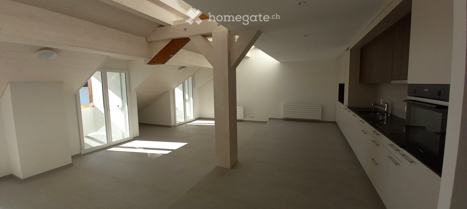 Open-plan space with a modern kitchen area featuring cabinets, a sink, and an oven, connected to a living area with large windows and wooden beams.