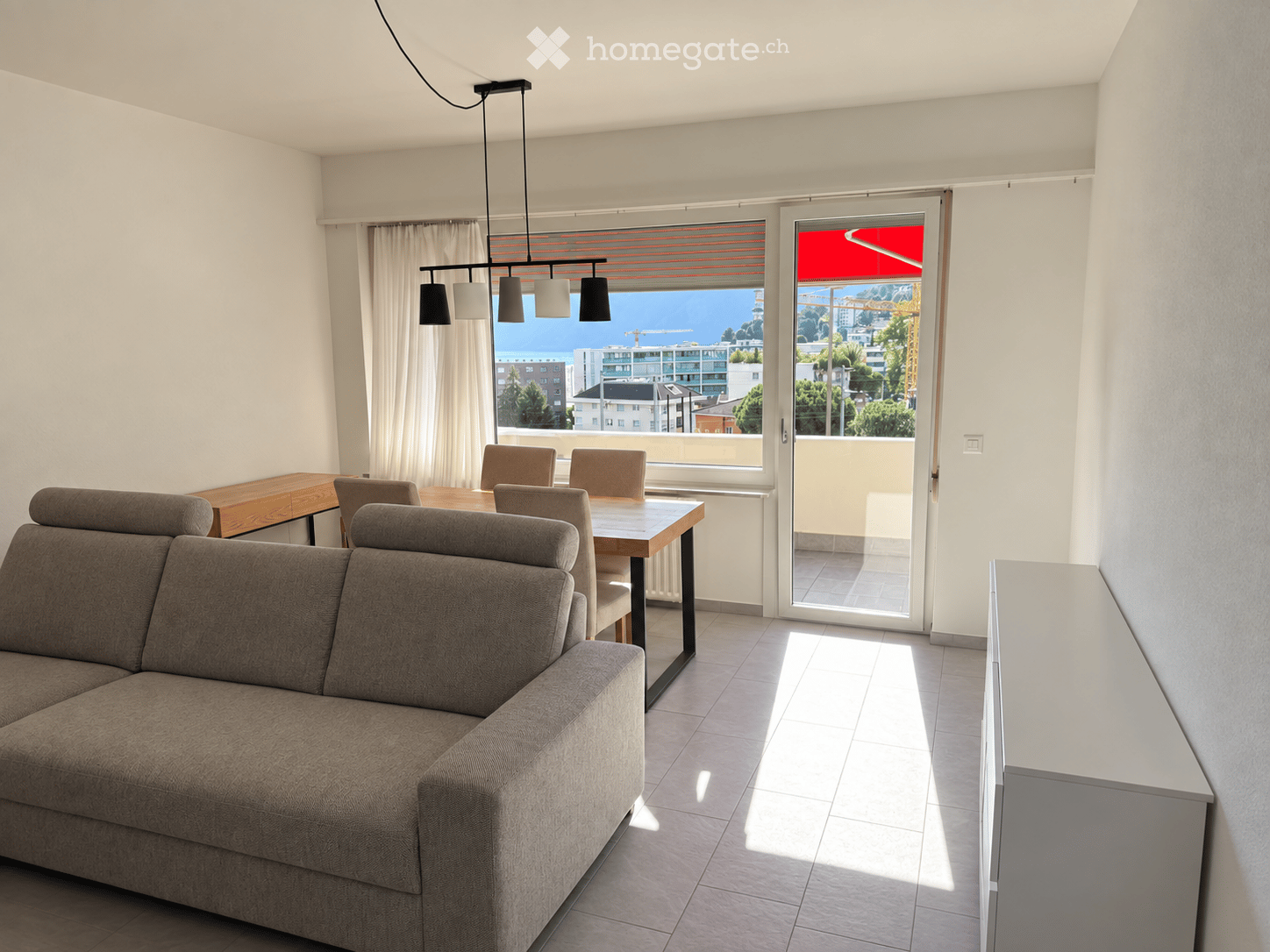 Modern living and dining area with a grey sectional sofa, wooden dining table, chairs, pendant lighting, and large windows offering a scenic view.
