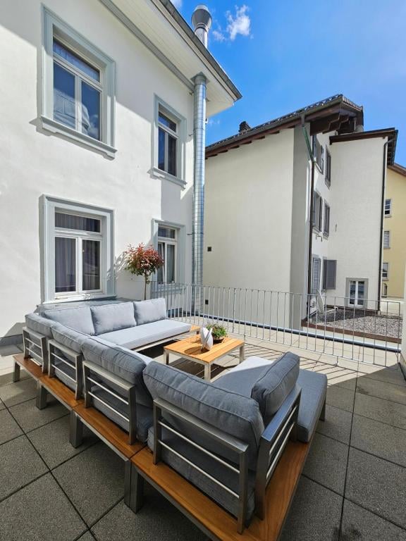 patio, outdoor seating, metal railing, wooden bench, coffee table, plant in vase, white brick building, window with curtain