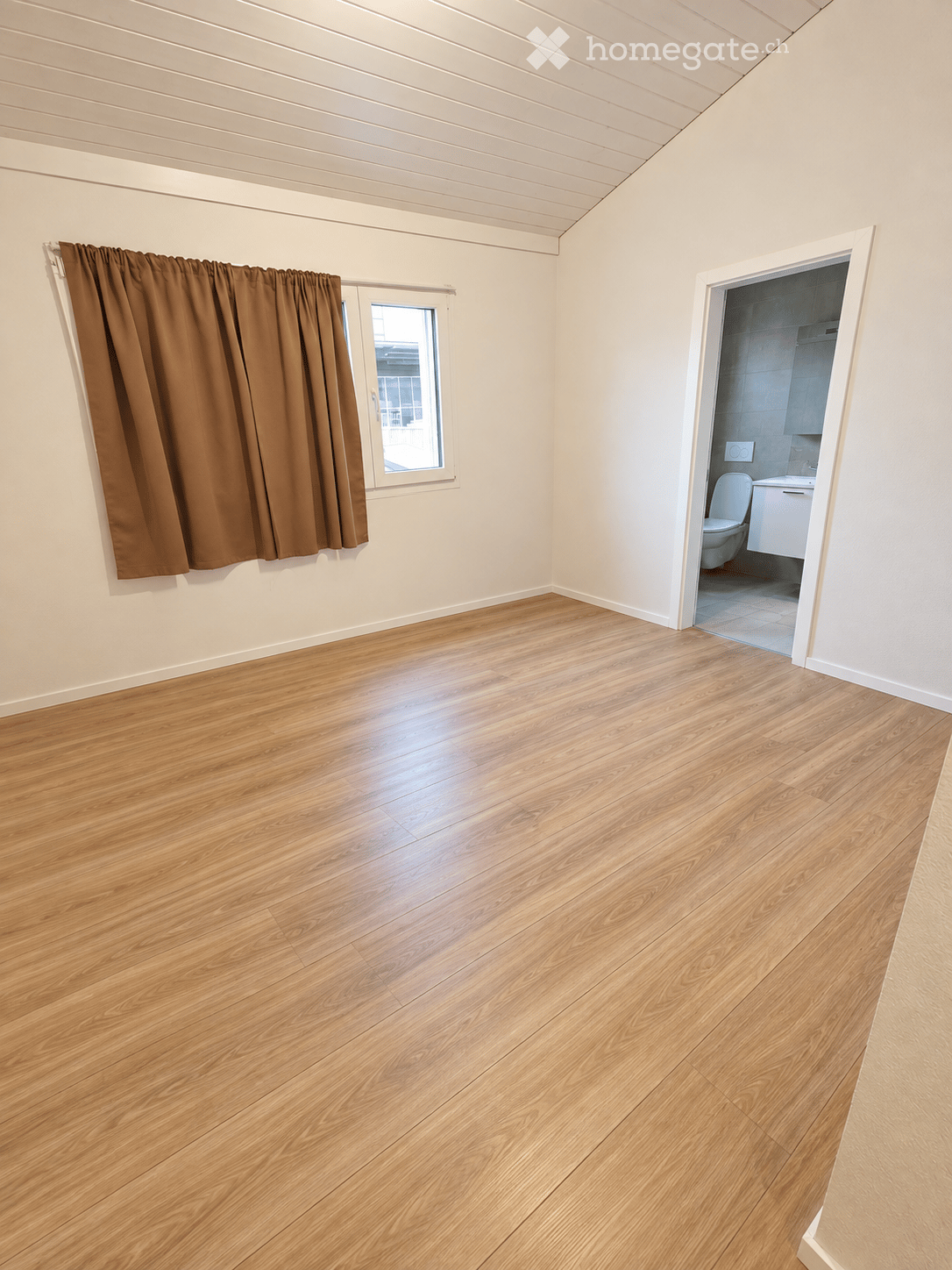Empty room with wooden flooring, white walls, and a window with brown curtains. An adjacent bathroom is visible through an open doorway.
