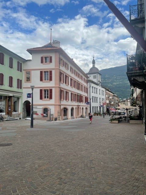 A charming European town square with a multi-story building featuring red shutters and a dome-shaped roof, surrounded by other historic buildings and a scenic mountain backdrop.