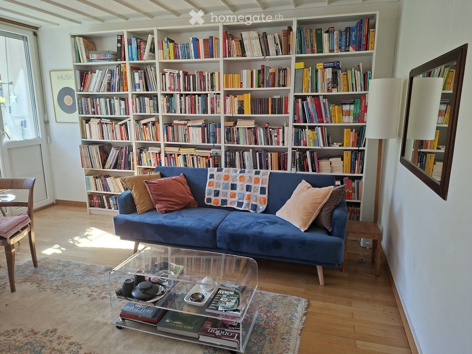 A cozy living room with a blue sofa, throw pillows, a glass coffee table, a bookshelf filled with books, a wooden chair, and a floor lamp.