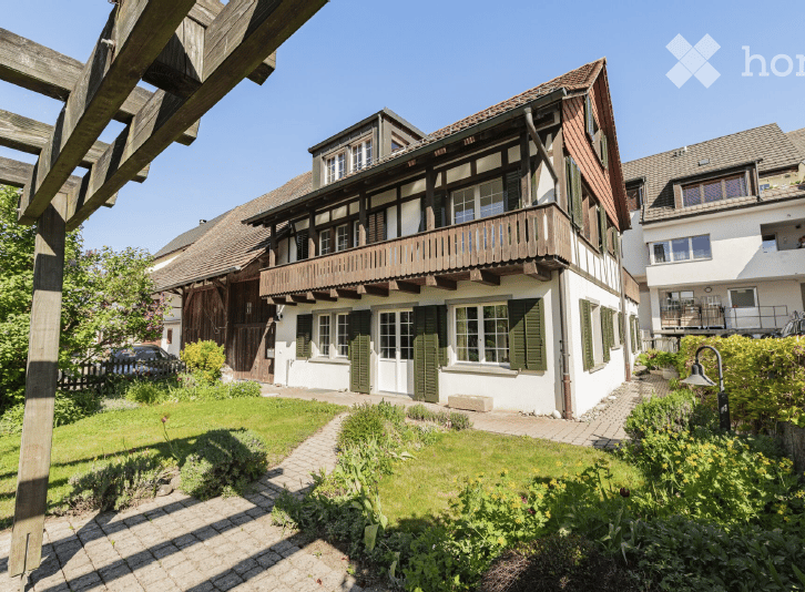Traditional two-story house with white walls, green shutters, and a wooden balcony. The house features a well-maintained garden with a paved pathway leading to the entrance.