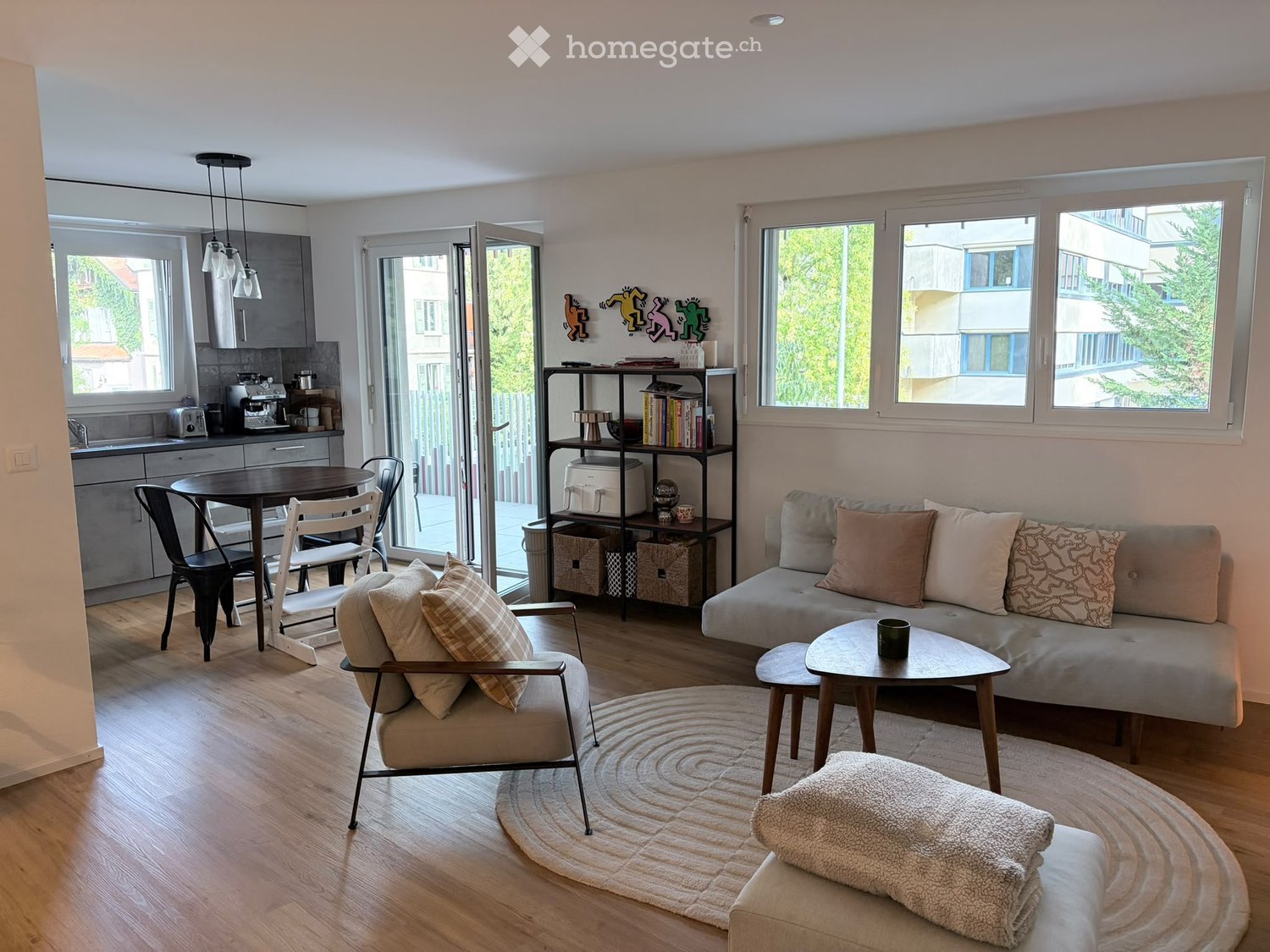 A modern, open-concept living and dining area with a sofa, coffee table, armchair, dining table, and kitchen area. The space features large windows, wooden flooring, and decorative pillows on the sofa.