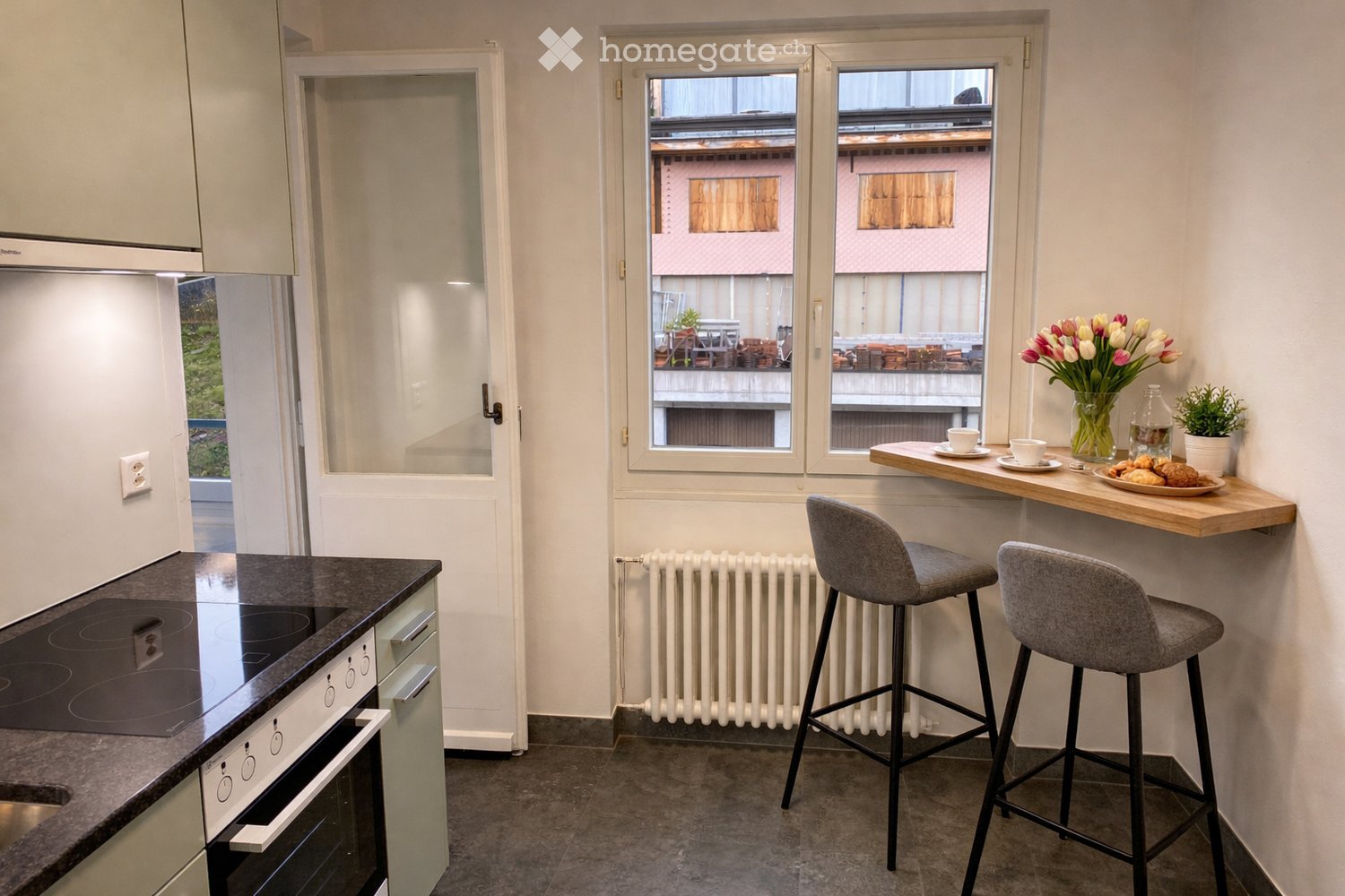 Modern kitchen with an induction stove, oven, bar counter, and two bar stools. A vase with tulips, a cup, a bottle, and a plate with pastries are on the counter. A potted plant is also visible.