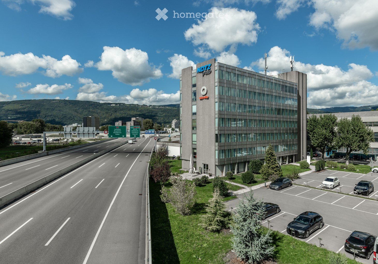 A multi-story office building with the Notz logo on the facade, surrounded by a parking lot and a highway with multiple lanes of traffic.