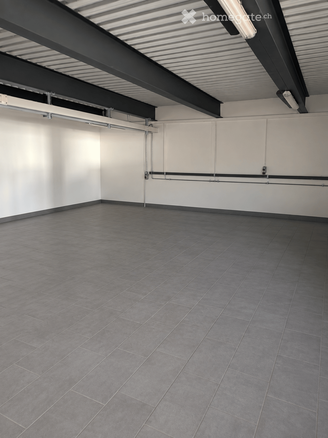 Empty room with grey tiled floor and white walls, featuring exposed metal beams and lighting fixtures on the ceiling.