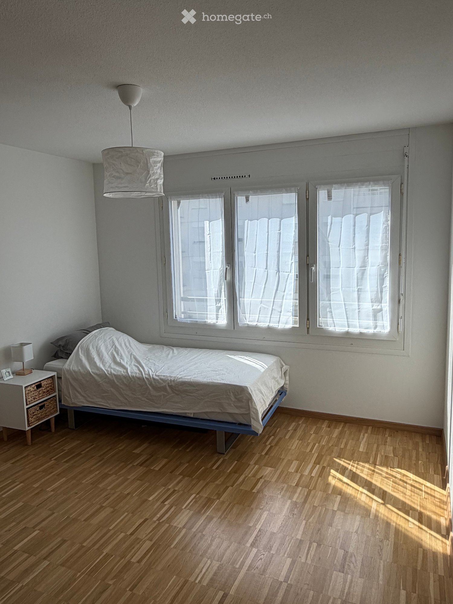 Minimalist bedroom with a single bed, nightstand, lamp, and picture frame. Large windows with sheer curtains allow natural light to fill the space.