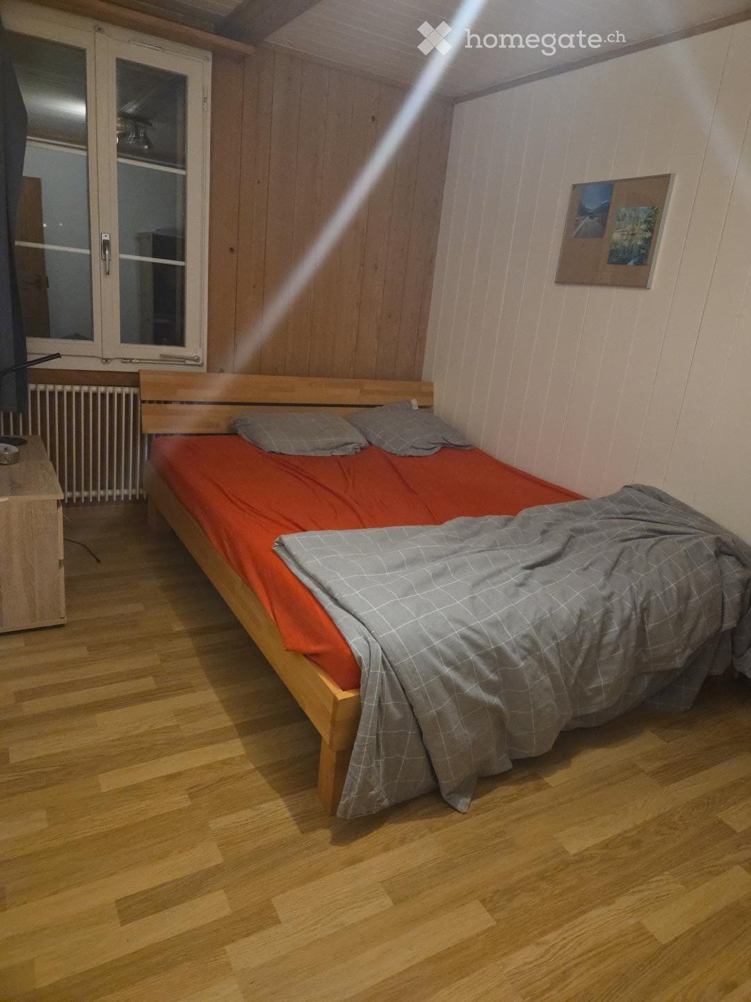 Cozy bedroom with wooden flooring, a single bed with red and grey bedding, a nightstand, radiator, and a picture frame on the wall.