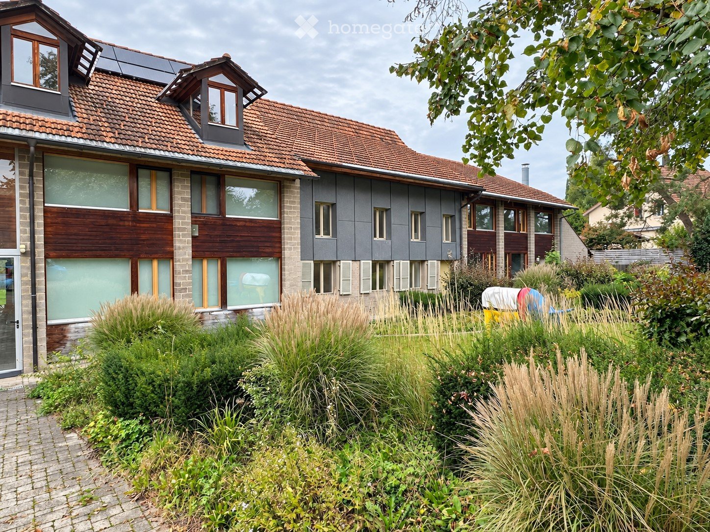 Modern two-story house with red-tiled roof, large windows, and solar panels on the roof. The house features a mix of brick and wood exterior, surrounded by a well-maintained garden with tall grasses and shrubs.